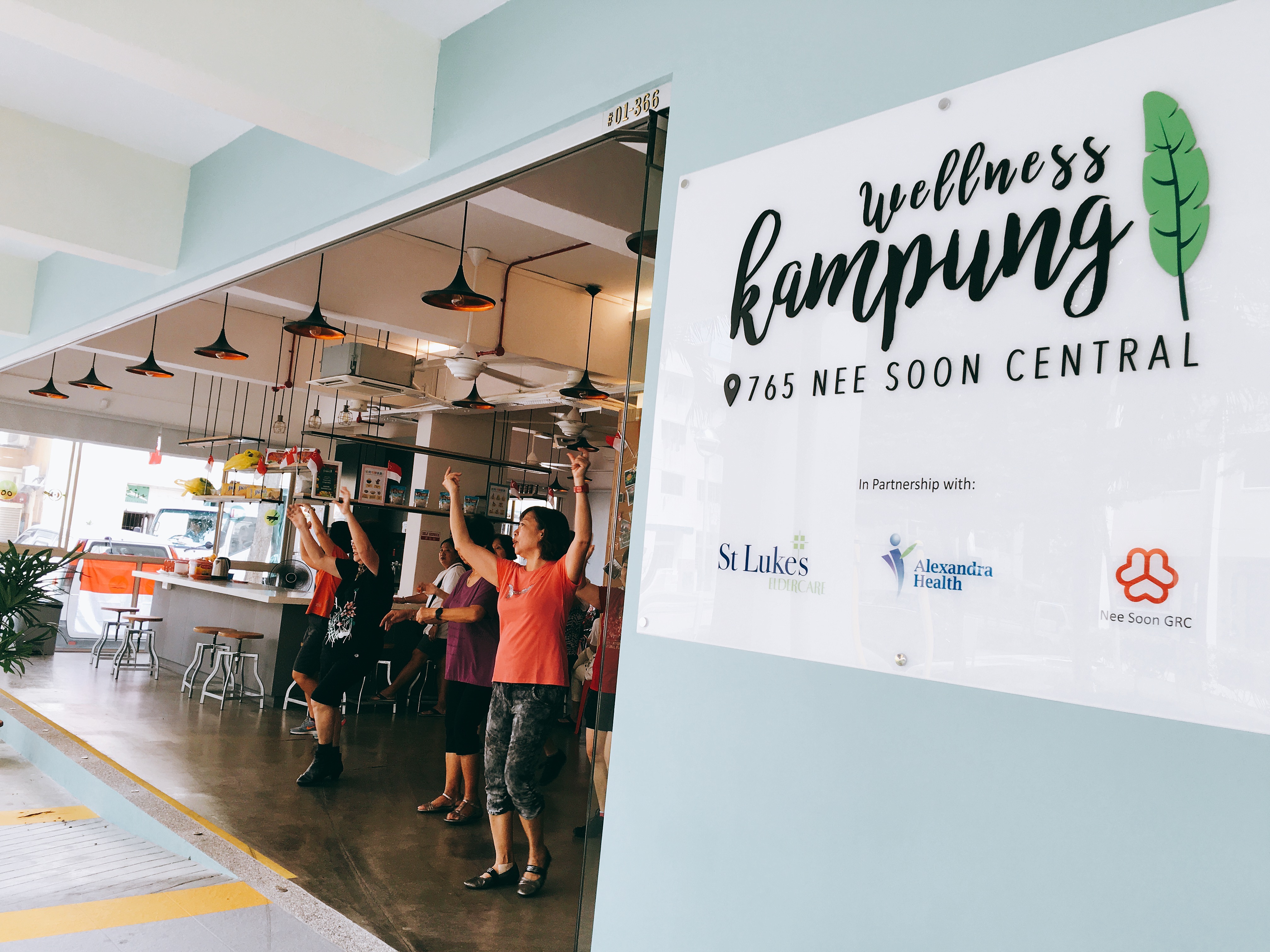 A group of residents exercise at the Wellness Kampung. The Wellness Kampung logo is prominently shown